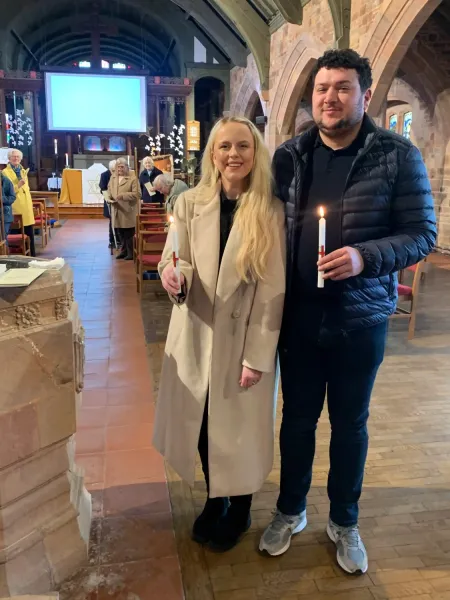 Jack and Georgie standing by the font in St. Sabinus' church with members of the congregation in the background.  Both are holding lit baptism candles